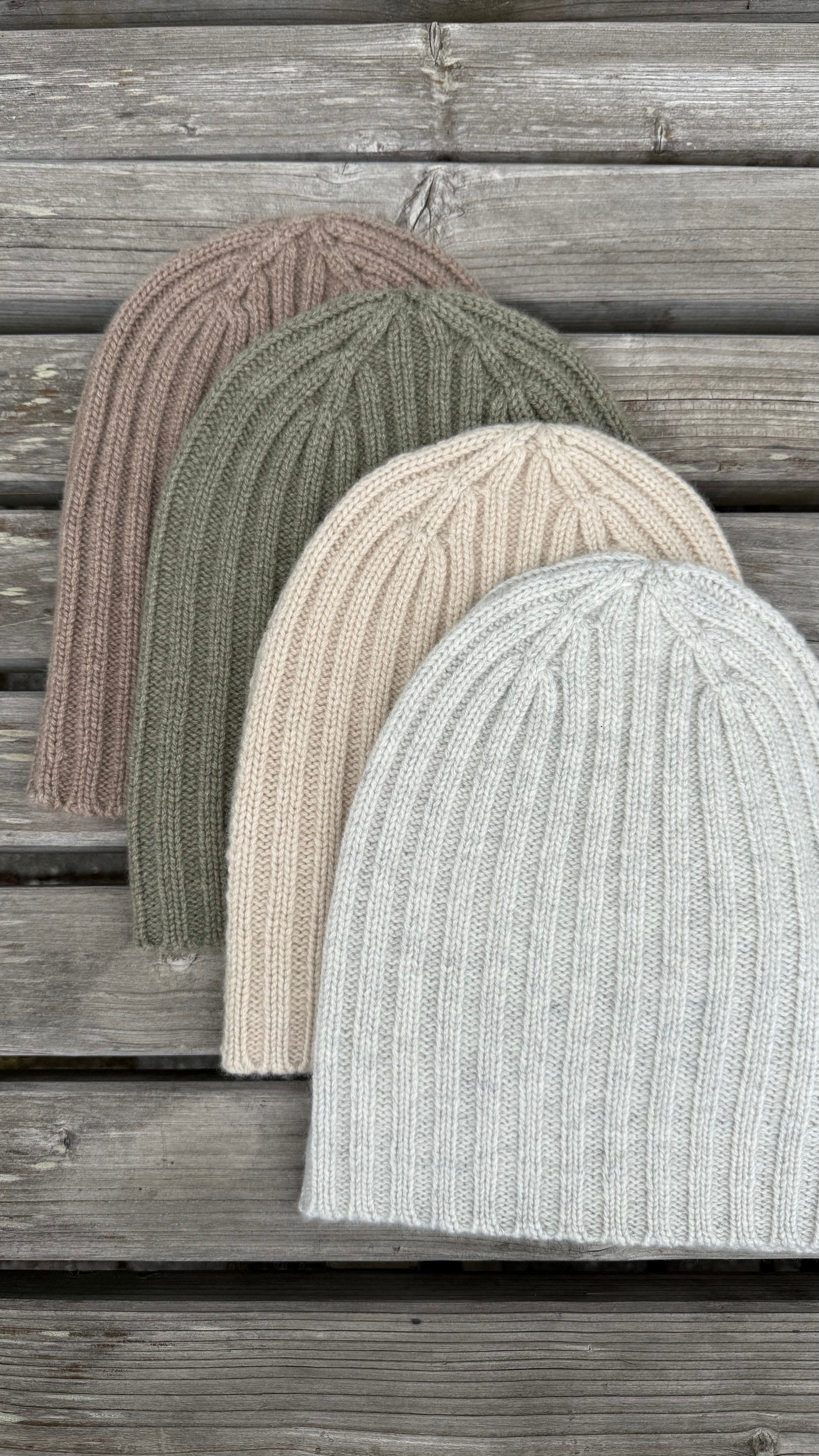 Four knit beanie hats in brown, green, undyed, and gray on a wooden surface.